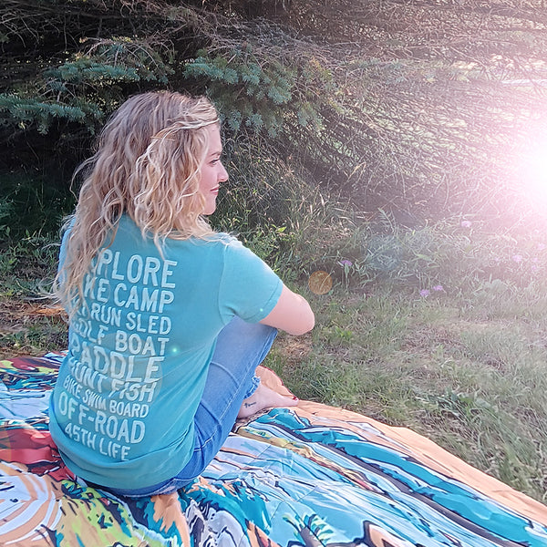 Woman wearing 45th Parallel Puff Ink Activity Tee in Sea Foam Blue, sitting outdoors on a colorful blanket with trees and sunlight in the background