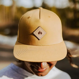 Tan flat-brimmed hat with leather patch featuring three tree design worn outdoors.