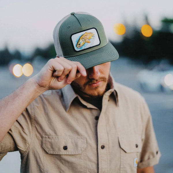 Man wearing 45th Parallel Trout Patch Trucker hat adjusting hat outdoors