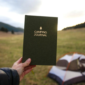 45th Parallel Basecamp Journal held up outdoors with tent and open field in background