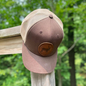 Brown 45th Parallel snapback trucker hat with elk leather patch, resting on wooden post, surrounded by green foliage